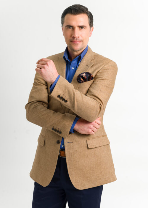 A men's tan linen herringbone jacket, styled with dark blue shirt and pocket square.